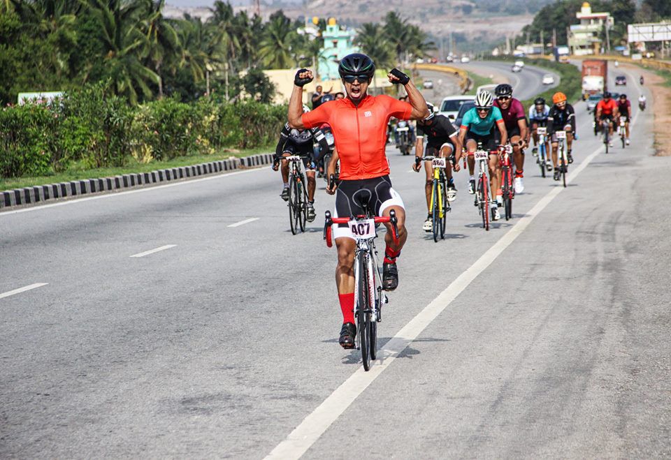 A BBCh rider celebrates crossing the line, arms raised, with the peloton behind on an open highway